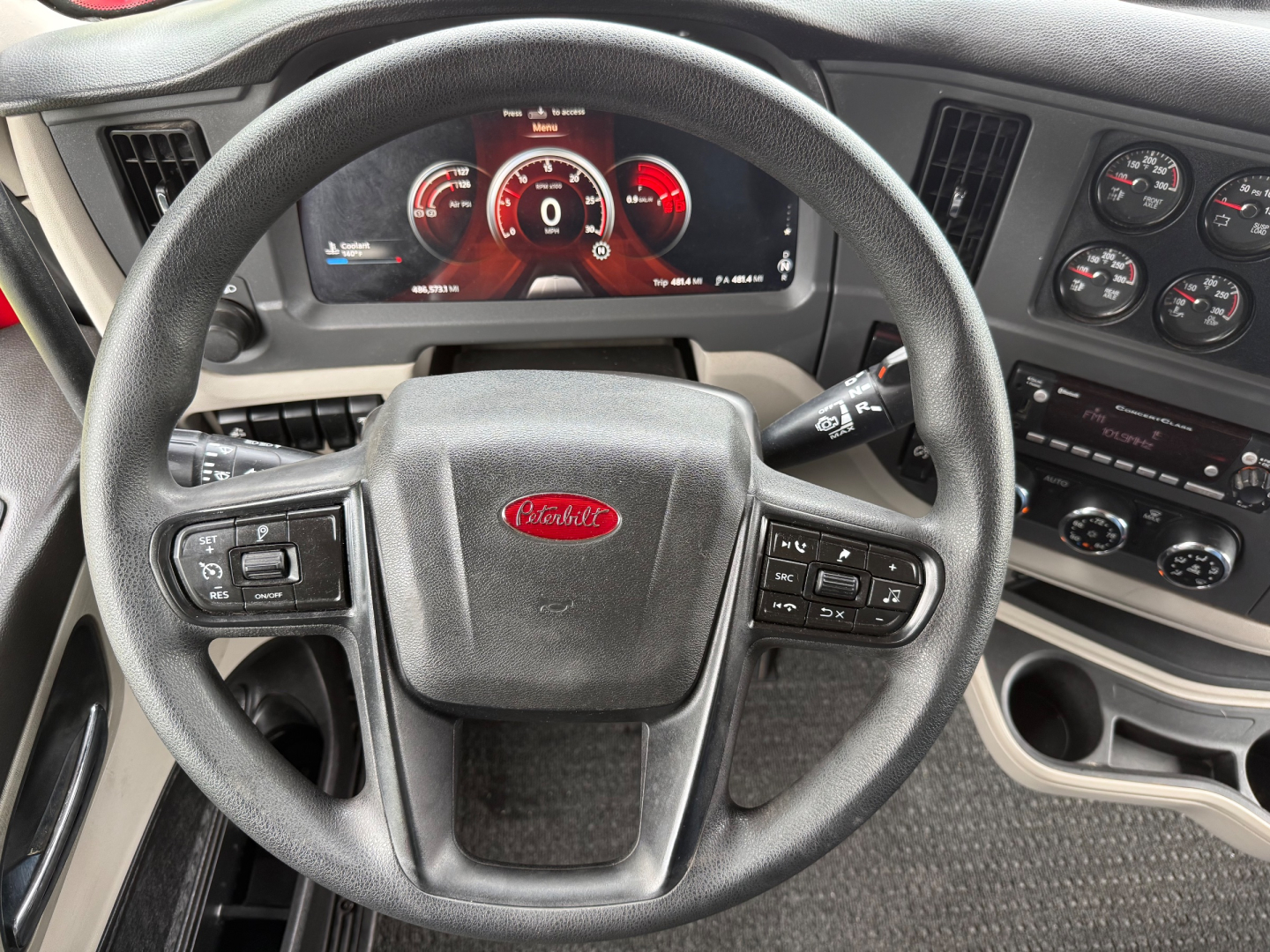 Peterbilt 579 interior steering wheel with digital gauge cluster showing red accents; steering wheel center badge Peterbilt; dashboard controls and gauges visible.