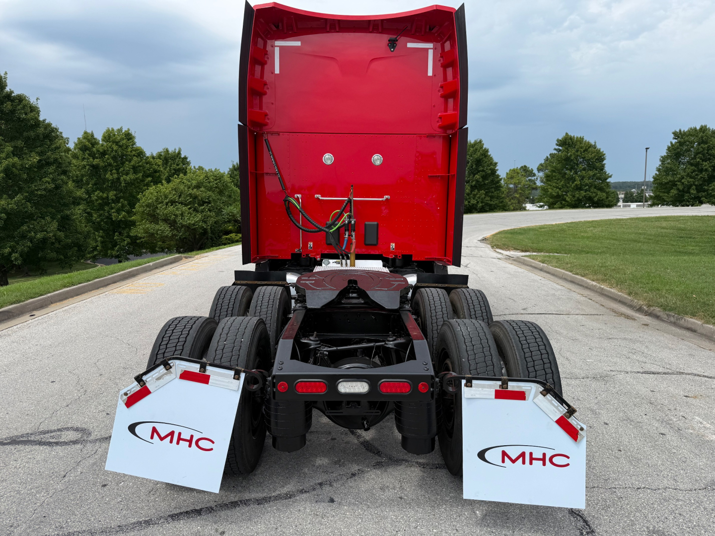 Peterbilt 579 sleeper tractor rear view showing fifth wheel and dual rear axles, red cab back, white mud flaps with MHC logo, road in background.