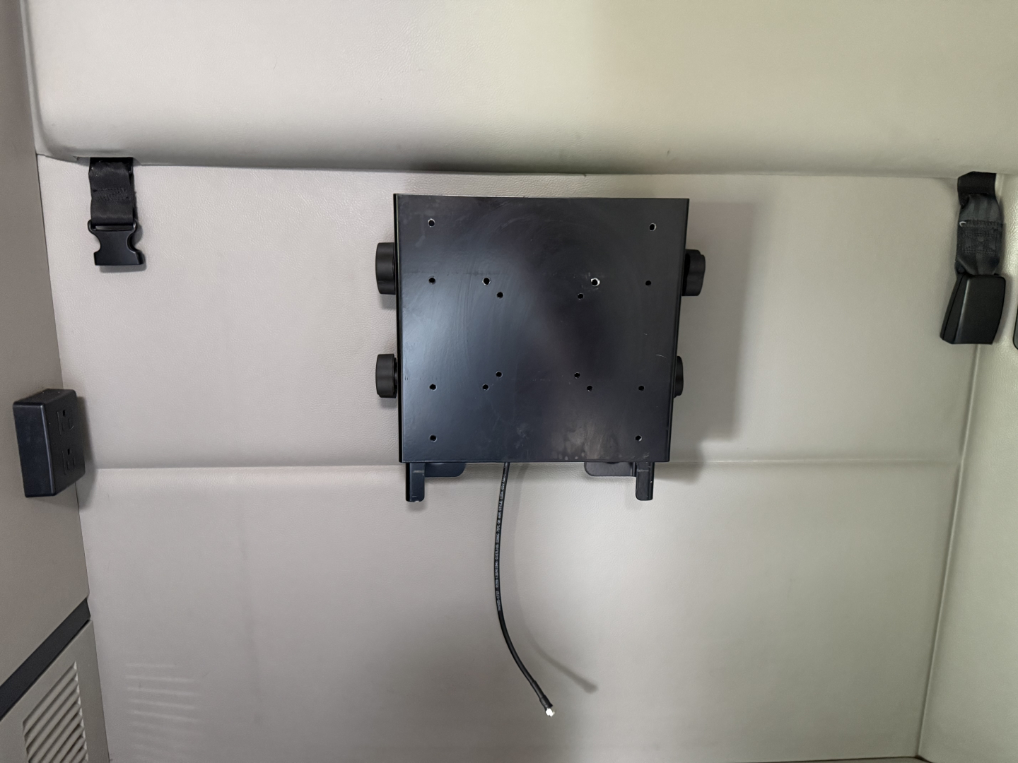 Vehicle interior panel showing a mounted black rectangular device with a coiled cable hanging down, on a light gray wall with latch hooks.