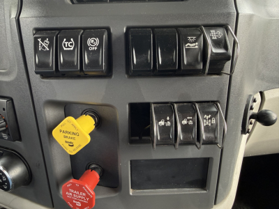 Peterbilt 579 dash cluster showing multiple switch panels and yellow parking brake knob; visible stickers read parking brake and trailer air supply; on the left a red trailer air supply knob; various icon buttons with OFF and other symbols.