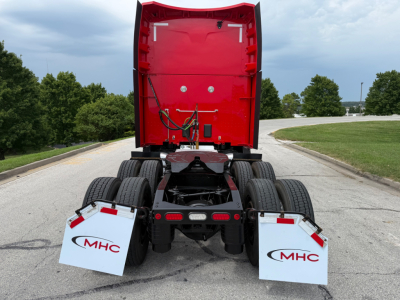 Peterbilt 579 sleeper tractor rear view showing fifth wheel and dual rear axles, red cab back, white mud flaps with MHC logo, road in background.