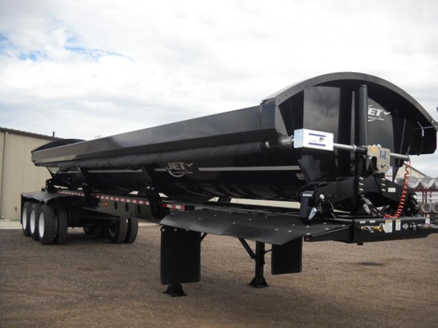 Tri-axle side dump trailer with black exterior, Jet brand, long rectangular hopper, landing gear down, front coupler and hoses visible.