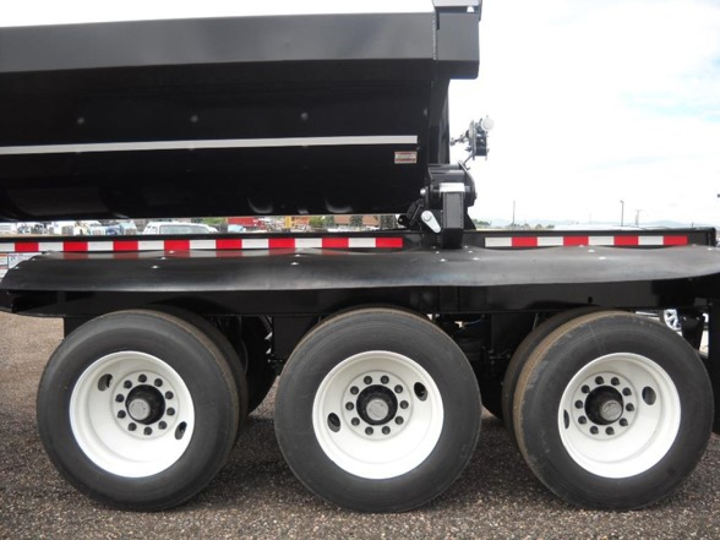 Trailer axle section showing three dual-tire wheels under a side-dump trailer with black dump bed and red/white reflective tape