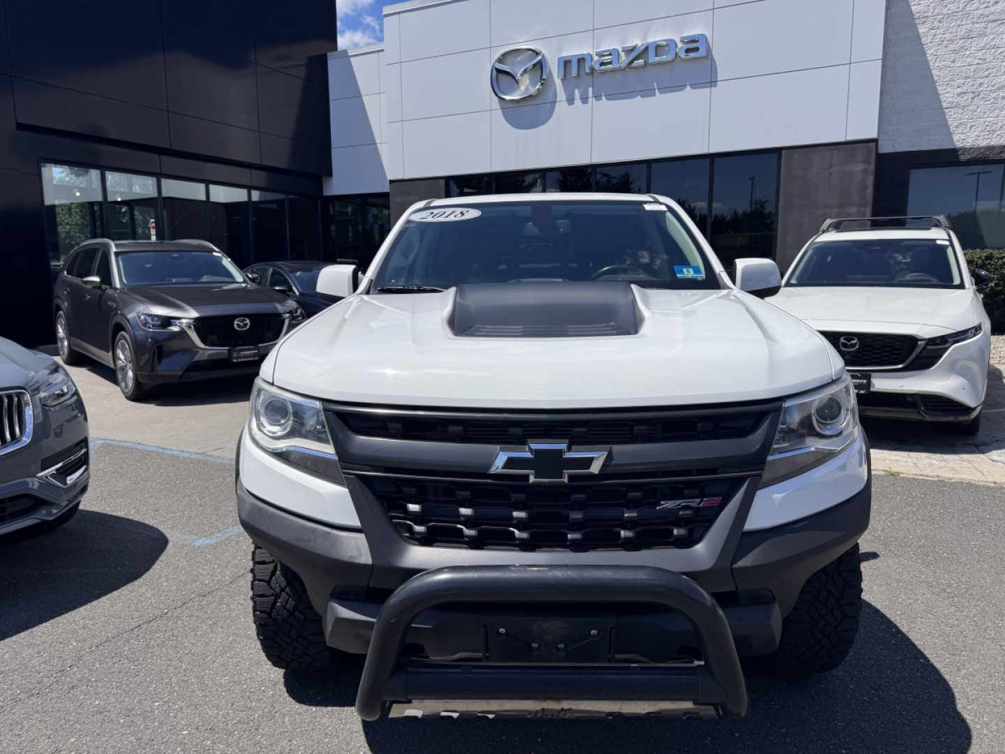 Front view of a white 2018 Chevrolet Colorado with aftermarket grille guard, parked in a Mazda dealership lot among other vehicles.