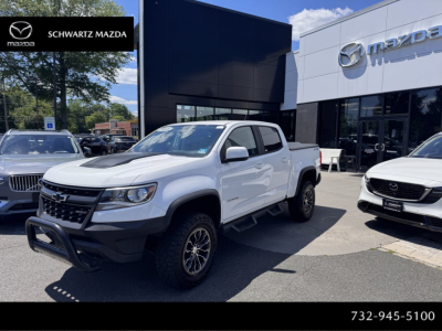 2018 Chevrolet Colorado crew cab pickup truck in dealership lot with white body, black bumper guard, and chrome wheels; dealership building and Mazda logo in background.