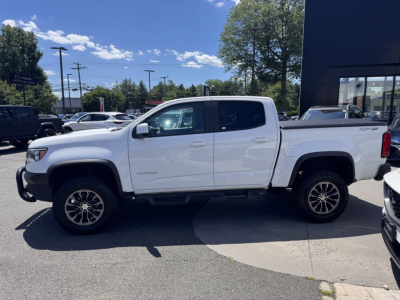 2018 Chevrolet Colorado double cab pickup truck in white viewed from the side, standing on a dealership lot.