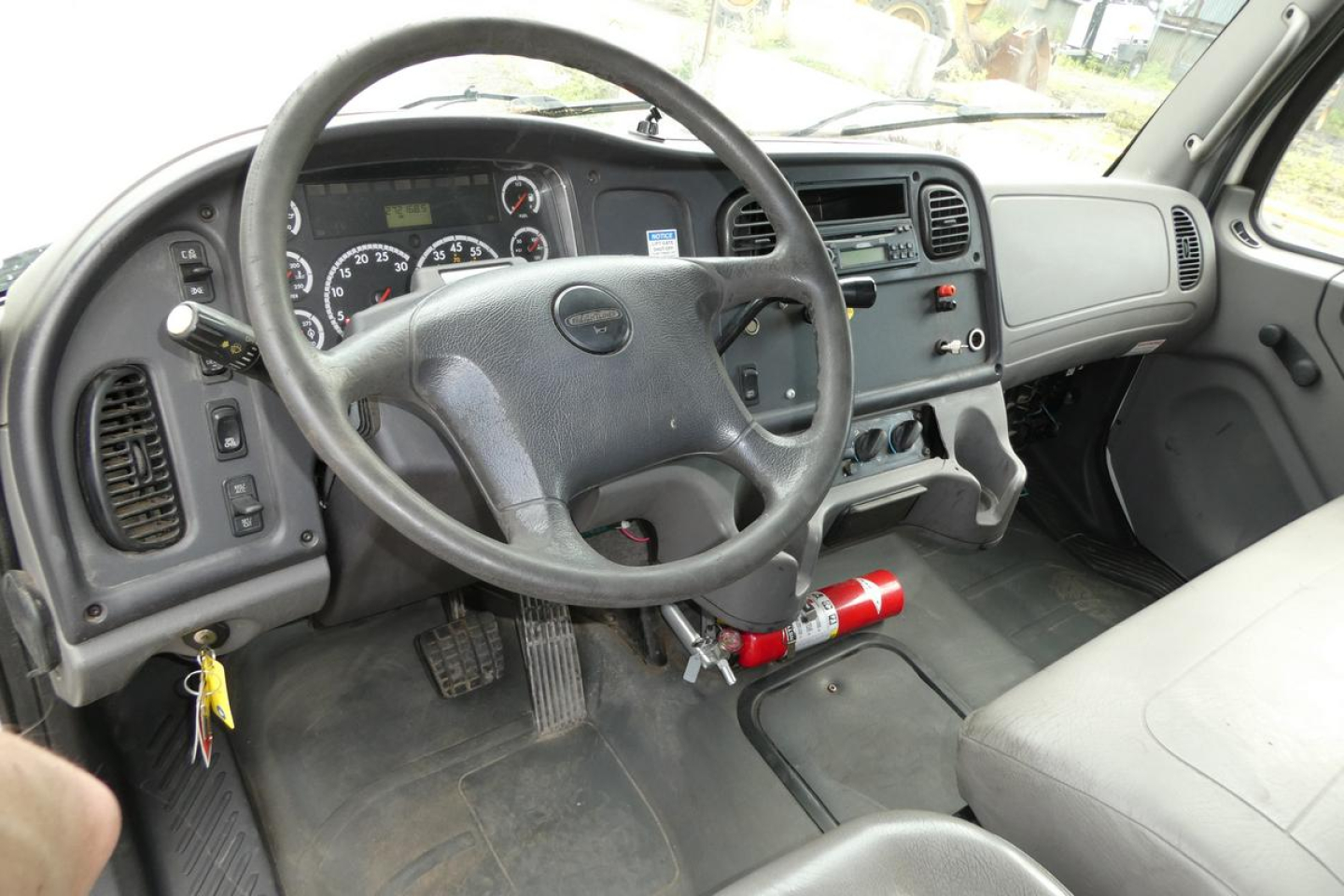 Cab interior of a Freightliner M2 106 truck showing steering wheel, dashboard gauges, gear shifter, and center console with a fire extinguisher mounted under the dashboard.