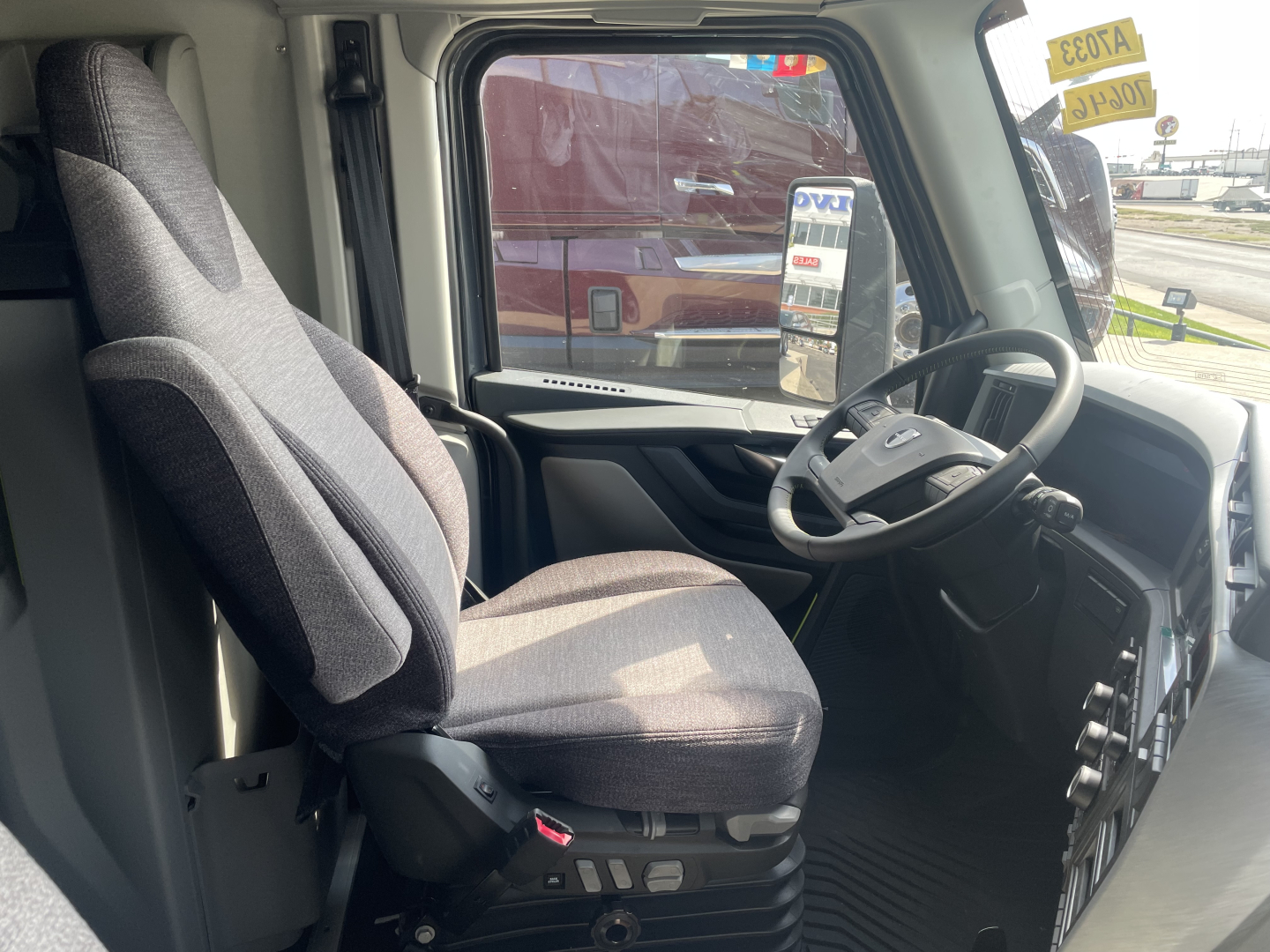 Truck cockpit interior with steering wheel, driver's seat, and dashboard; window shows exterior vehicles and dealership sticker.