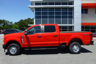 Side profile of a red 2026 Ford F-250 Super Duty pickup truck, extended cab, 4x4 badge visible, parked in front of a showroom.