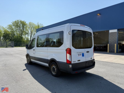 Ford Transit wheel chair van rear-right view showing back doors and wheelchair ramp area; white body with black bumper, large windows, blue sticker near rear, dealership building in background.