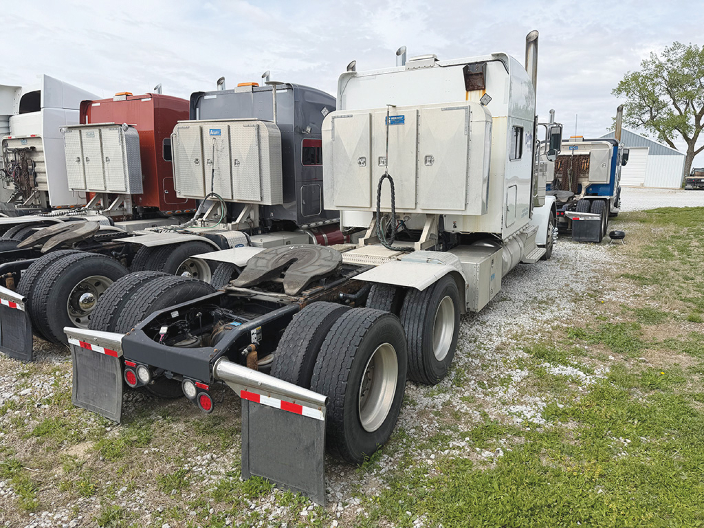 Truck tractor showing the rear chassis and toolboxes; white cab with dual rear axles and fifth wheel absent, fuel tanks and storage boxes mounted on sides, green grass and other trucks in background.