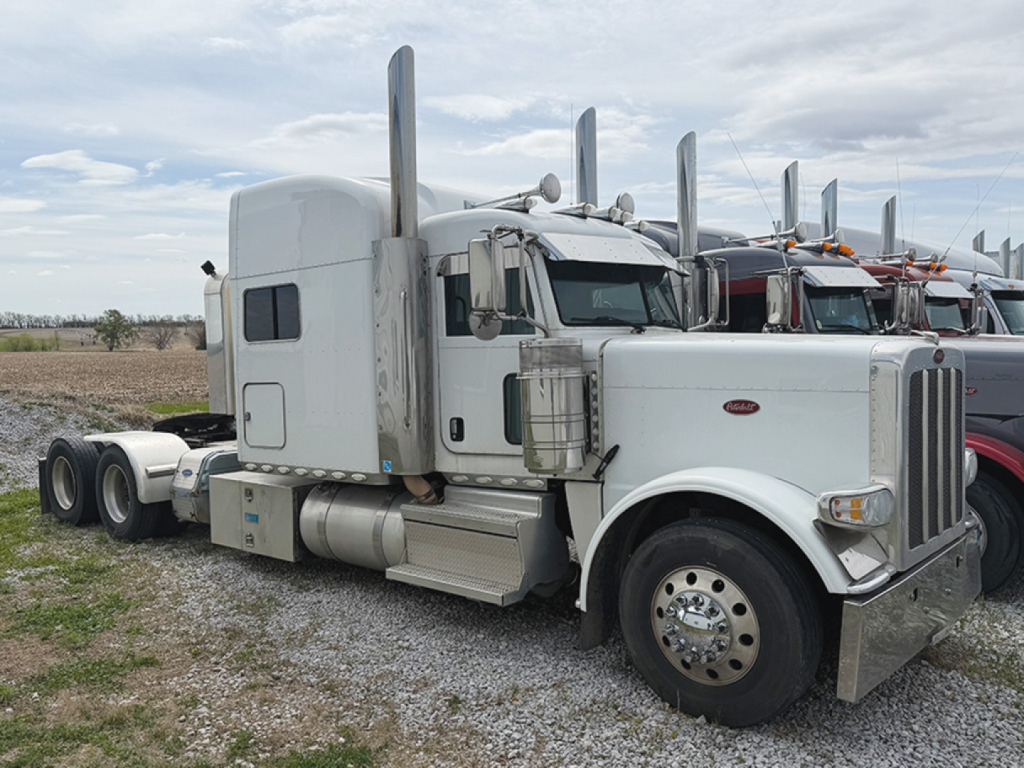 Peterbilt 389 UltraCab tag in a used truck lot showing a white conventional sleeper tractor with chrome exhaust stacks and dual rear axles.