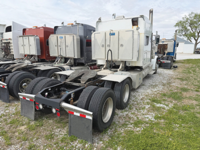 Truck tractor showing the rear chassis and toolboxes; white cab with dual rear axles and fifth wheel absent, fuel tanks and storage boxes mounted on sides, green grass and other trucks in background.