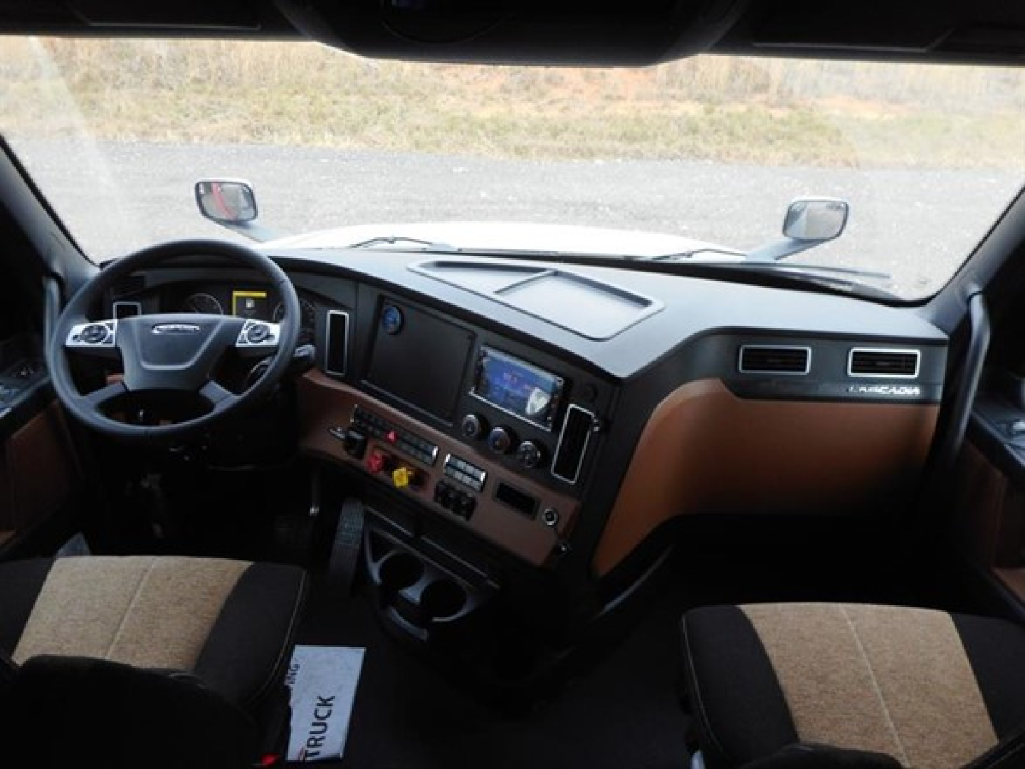 Dashboard view of a Freightliner Cascadia 126 cab interior showing steering wheel, dashboard controls, center touchscreen, and brown seat upholstery.