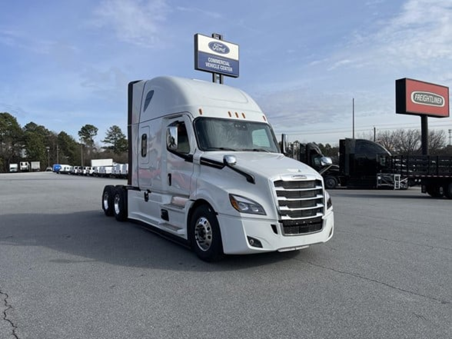 Freightliner Cascadia 126 tractor in a dealership lot, white cab with sleeper, chrome grille and bumper, several other trucks in background.