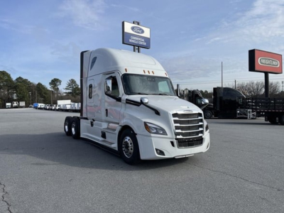 Freightliner Cascadia 126 tractor in a dealership lot, white cab with sleeper, chrome grille and bumper, several other trucks in background.