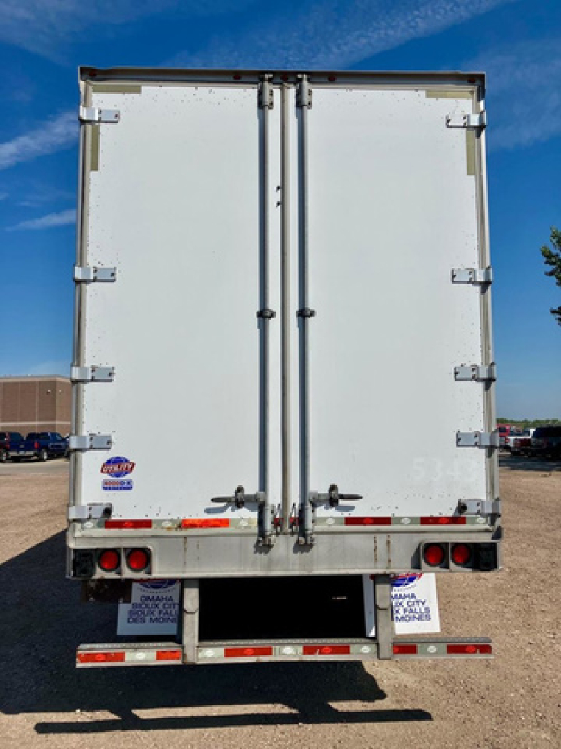 Rear view of a white utility van trailer with double doors and hinge hardware. VIN tag area and stock decals visible on lower rear bumper, with red and white reflective strips.
