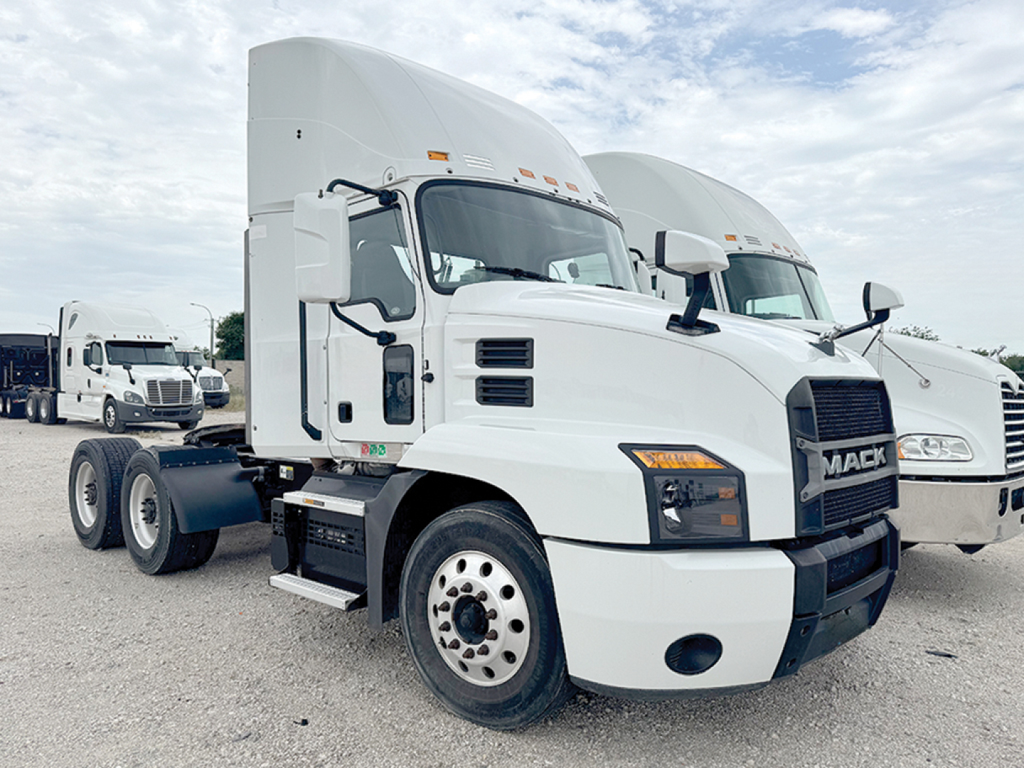 Mack Anthem 64T truck in white, side view showing cab and front with Mack grille, dual rear axles, and sleeper cab; outdoor lot with other trucks in background.