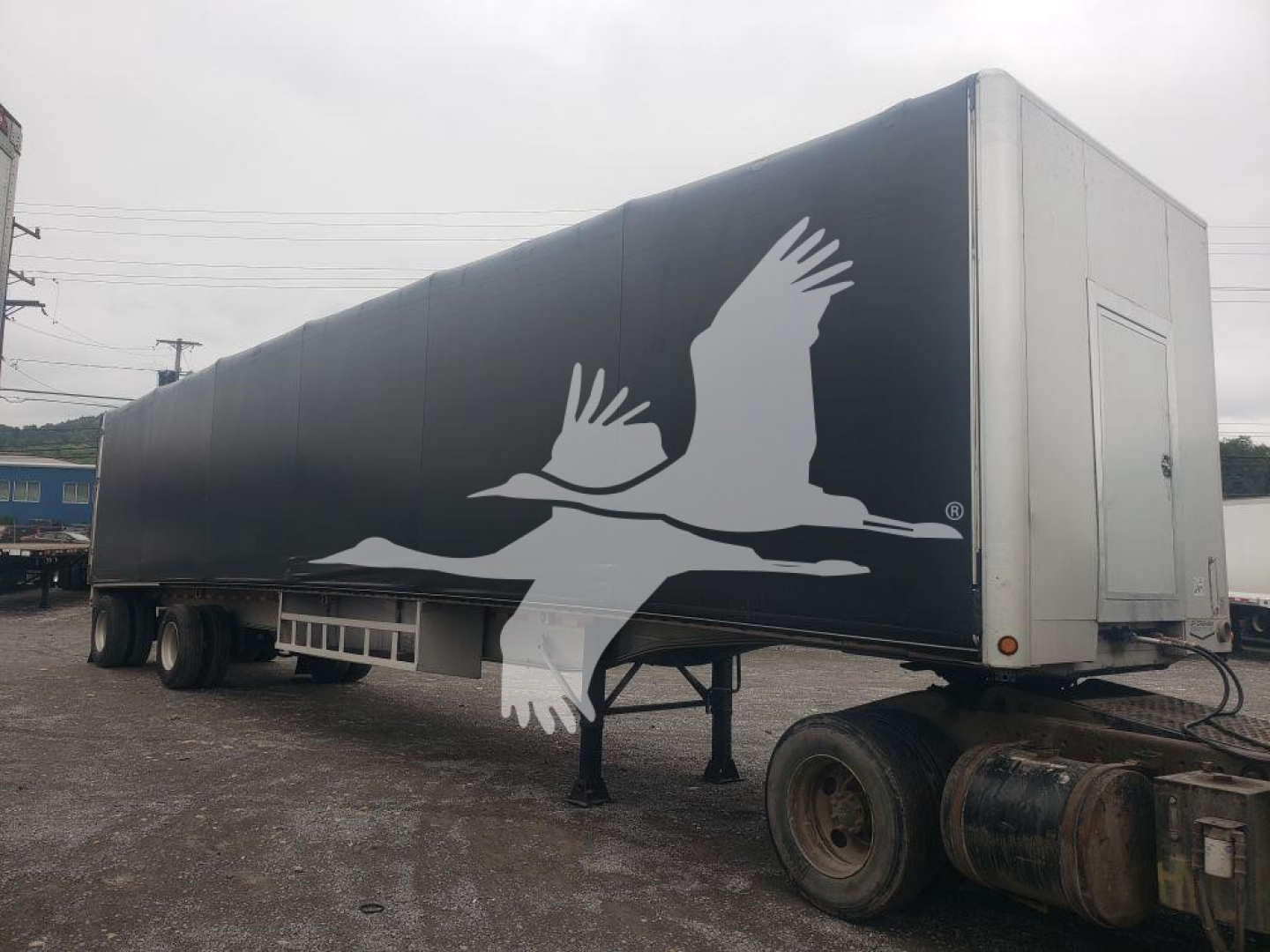 Flatbed trailer with curtainside body showing a white graphic of flying geese on dark gray side; front is silver with access door; wheel axles at rear.