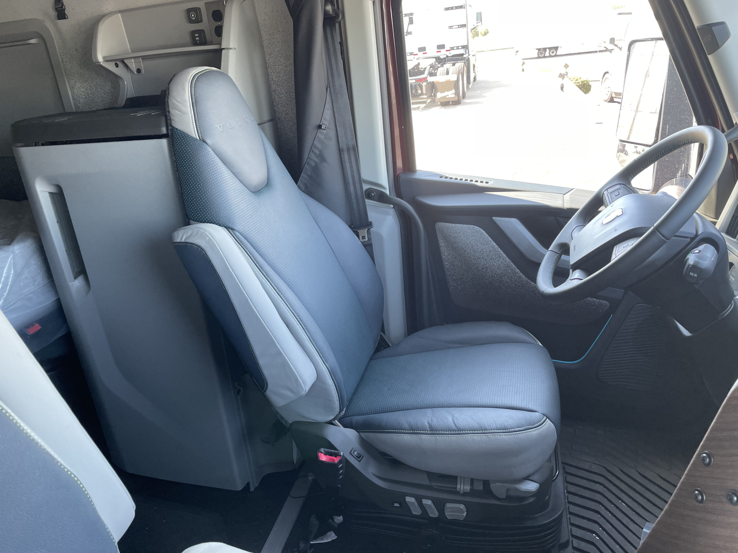 Volvo VNL cabin front interior showing driver seat and steering wheel with dashboard, light gray upholstery, and visibility of sleeper area.