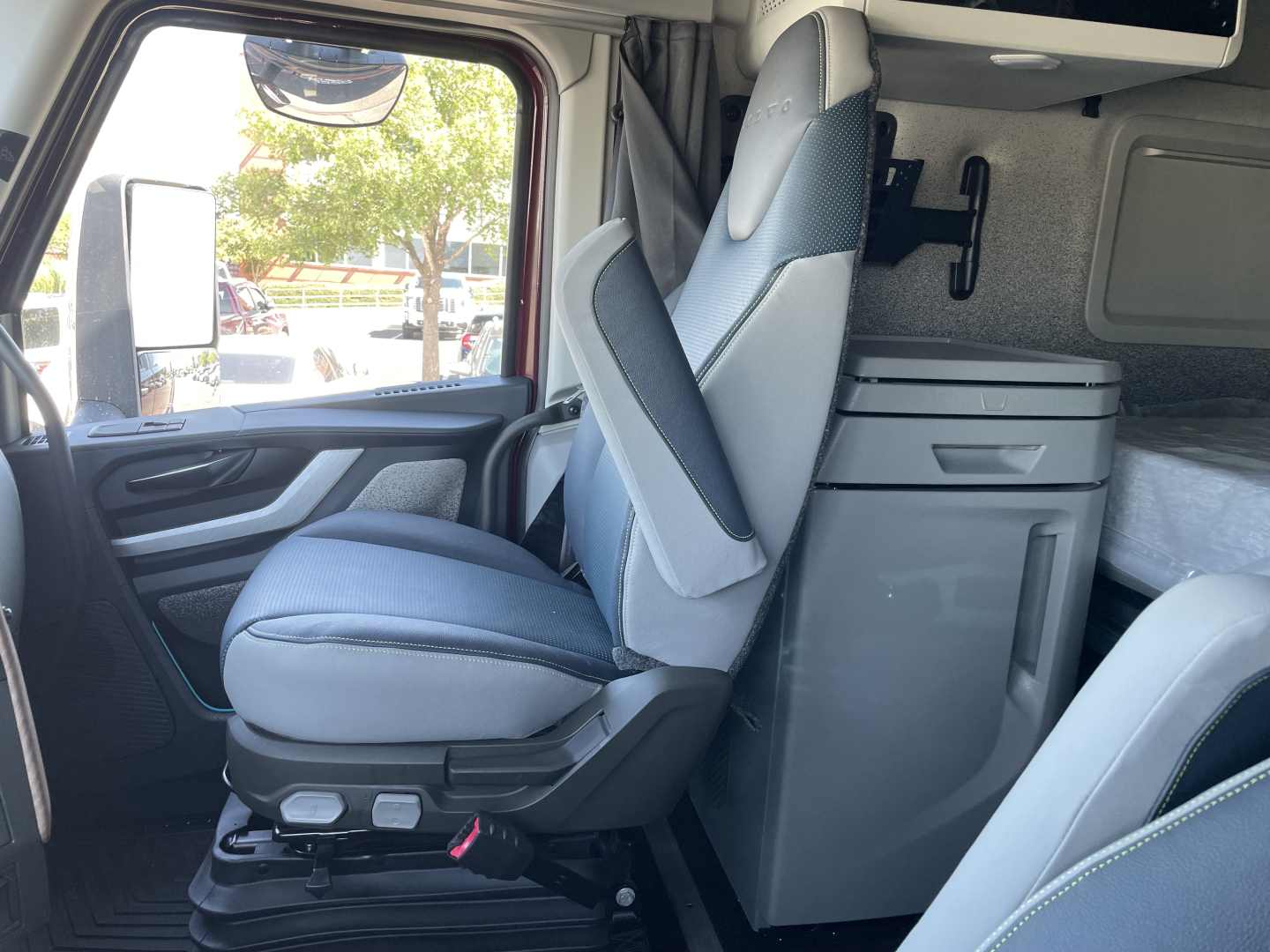 Volvo VNL64T860 cab interior with driver seat and living space; visible components include front seat with armrest, overhead sleeper cabinet, and compact refrigerator drawer unit.