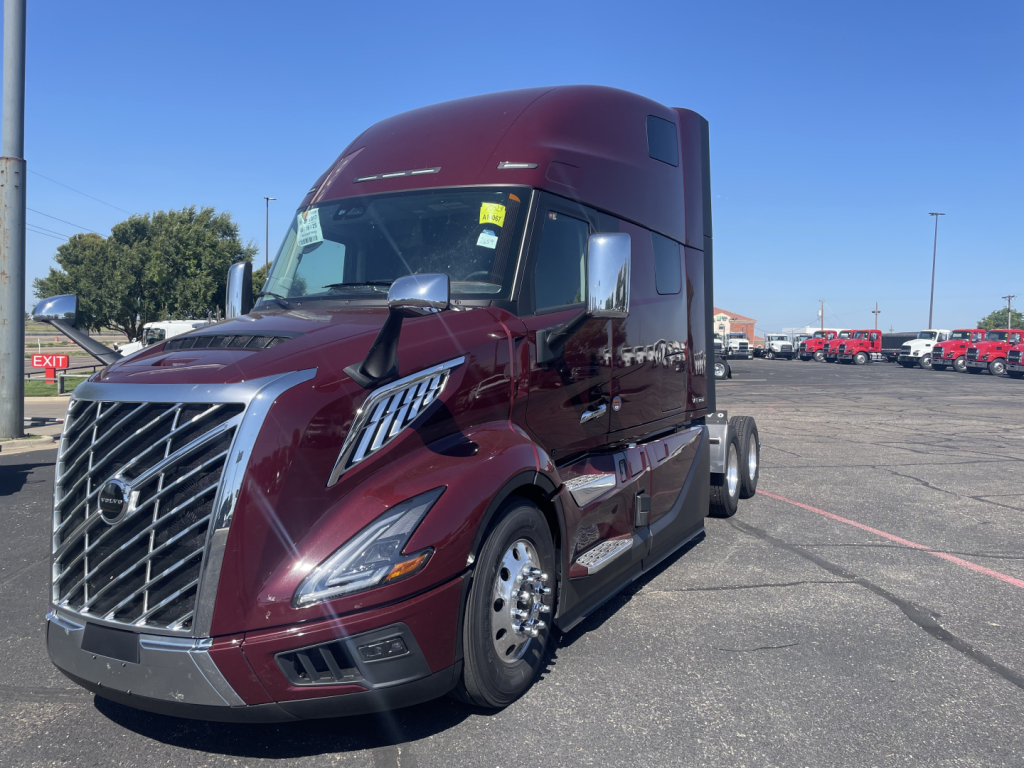 Volvo VNL64T860 tractor-trailer in burgundy showing front grille, cab with high roof, chrome mirrors, and dual rear wheels; daytime lot with other trucks in background.