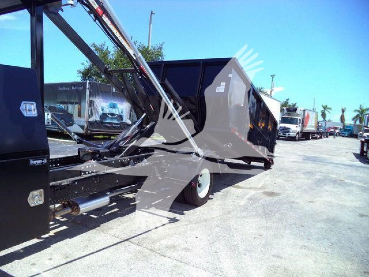 Isuzu roll-off truck body with hydraulic arms and dump bed, side view showing blue sky and other trucks in lot.