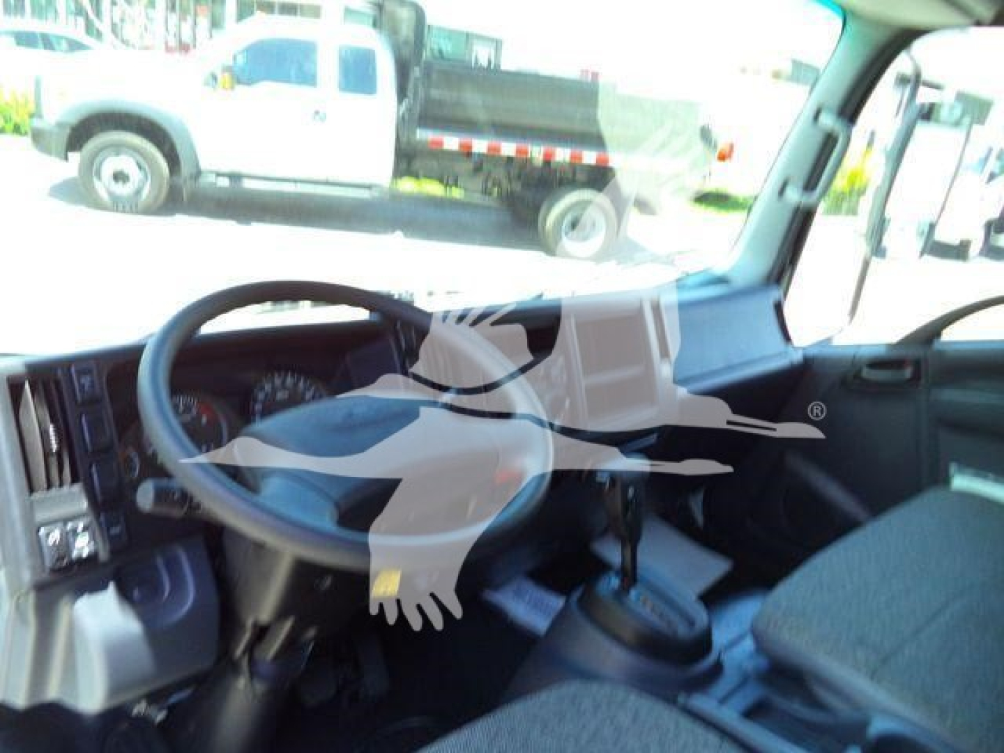 Isuzu NPRR truck cabin interior showing steering wheel, dashboard, and gear shift. Exterior work truck visible through windshield.