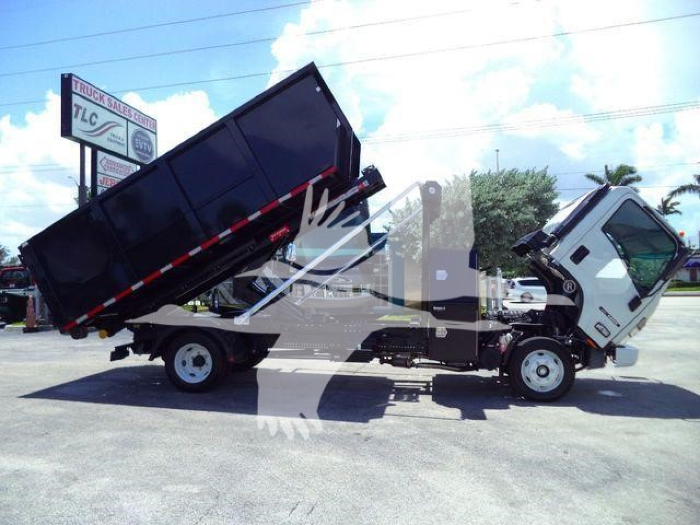 Isuzu NPRR roll-off truck with hydraulic bed partially raised, cab and chassis visible, in a dealership lot.