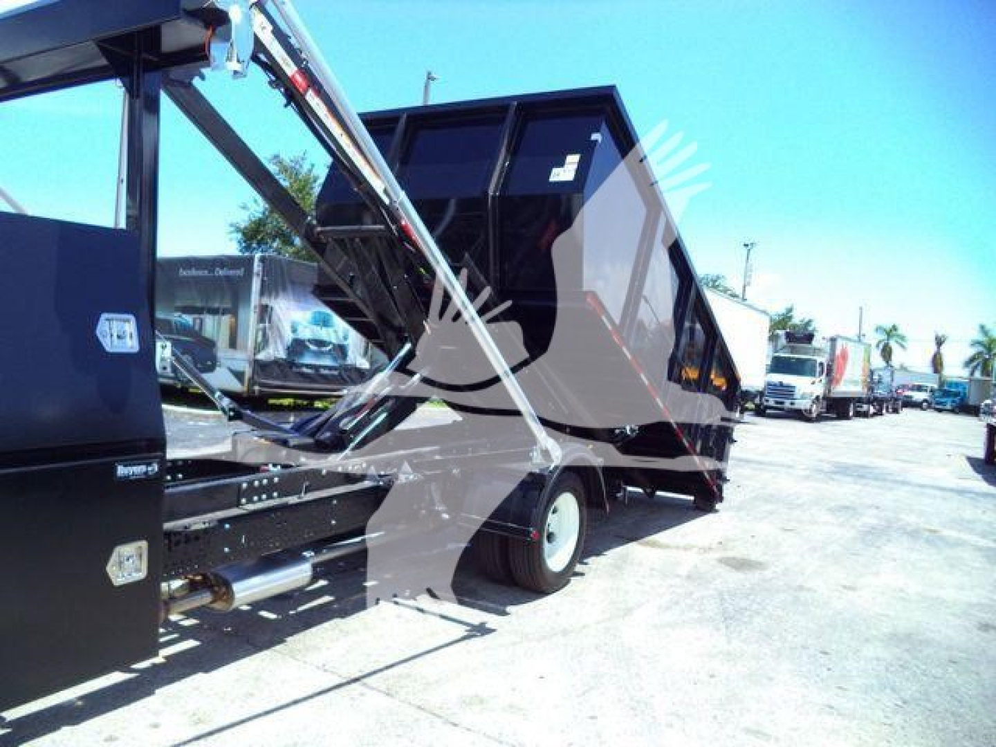 Isuzu NPRR roll-off truck bed in-service showing hydraulic arms and hopper in black with gray chassis and a row of trucks in the background.