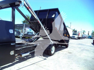 Isuzu NPRR roll-off truck bed in-service showing hydraulic arms and hopper in black with gray chassis and a row of trucks in the background.