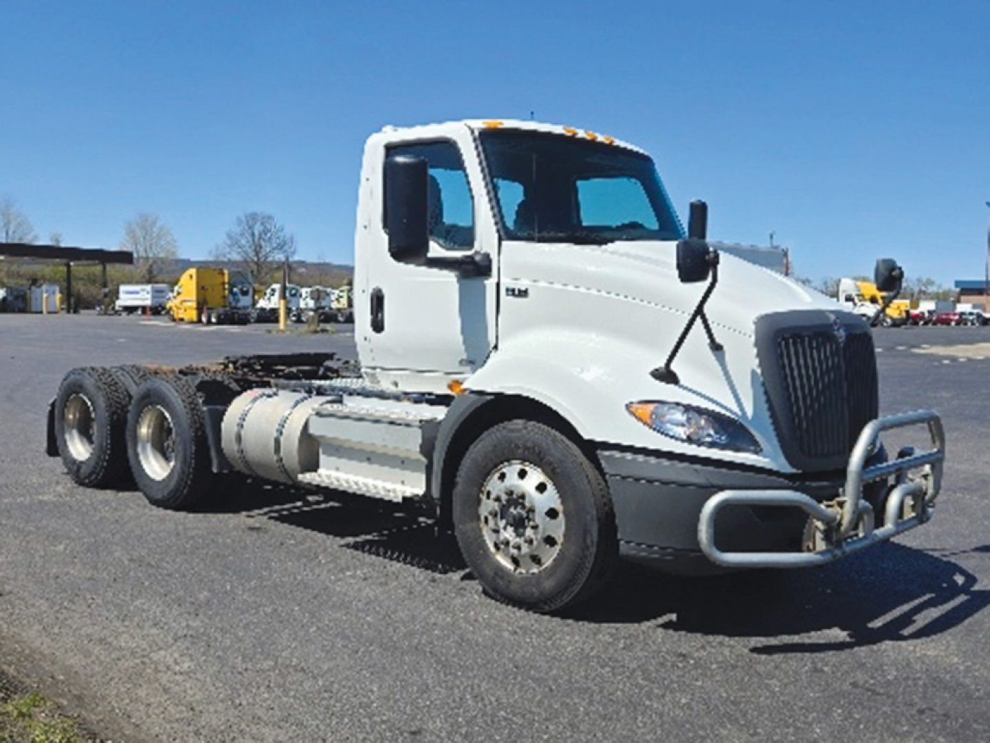 Truck cab and chassis showing International RH613 with front bumper and grille, 3-axle configuration, white cab, black tires, fuel tanks along side, warehouse lot in background.