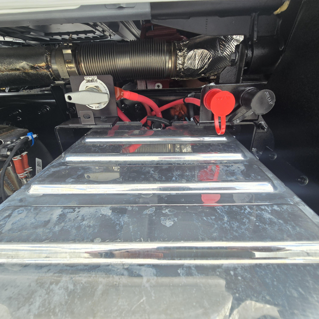 Mack PI64T engine bay showing battery with red and black cables, jump start terminal, air intake hose, and shiny metal fuel/air reservoir cover.