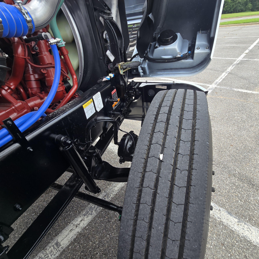 Truck front left undercarriage with large dual rear wheels, Mack PI64T engine bay visible, blue hoses and red engine components.
