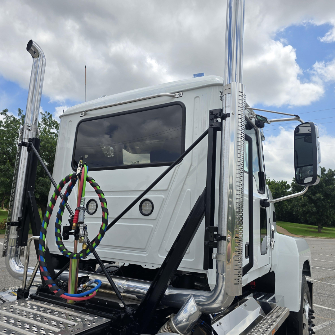 Truck cab showing air intake stacks and chrome exhausts with fuel lines and circular gauge on white Mack PI64T cab front.
