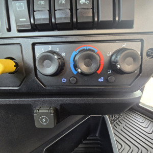 Cab cockpit controls of a 2027 Mack PI64T showing climate control dials (left fan speed, center temperature blue/red, right airflow) with 12V auxiliary power outlet below.