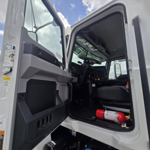 Cab interior of a 2027 Mack PI64T daycab truck with open door, dashboard controls, steering wheel, and emergency fire extinguisher visible.