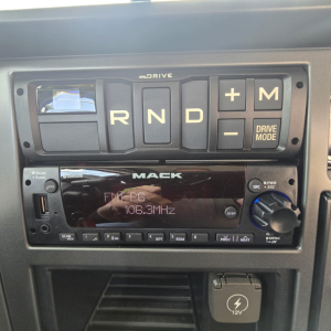 Mack PI64T dash cluster showing gear selector R N D plus M with drive mode; FM/AM radio unit displaying 106.3MHz; 12V power outlet symbol in lower console.
