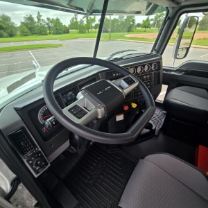 Interior cab view of a 2027 Mack PI64T, steering wheel, dashboard with multiple gauges, pedals, and driver seat visible.