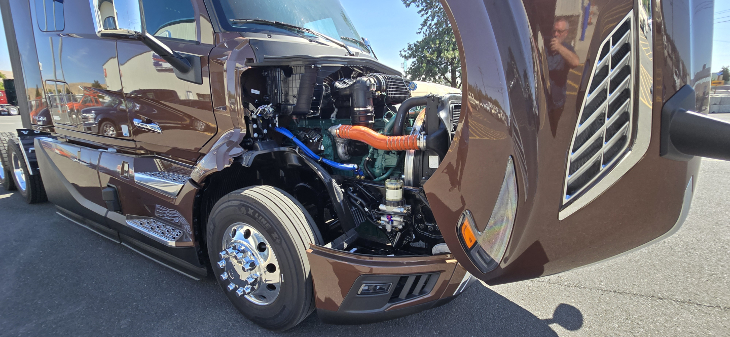 Volvo VNL64T860 front-left engine bay exposed showing blue hoses, orange coolant hose, chrome wheel, brown exterior, and open hood.