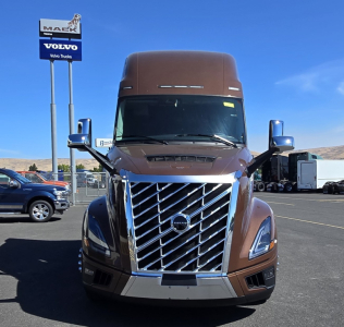 Front view of a brown Volvo VNL64T860 tractor-trailer with a large chrome grille and blue Volvo sign in background.