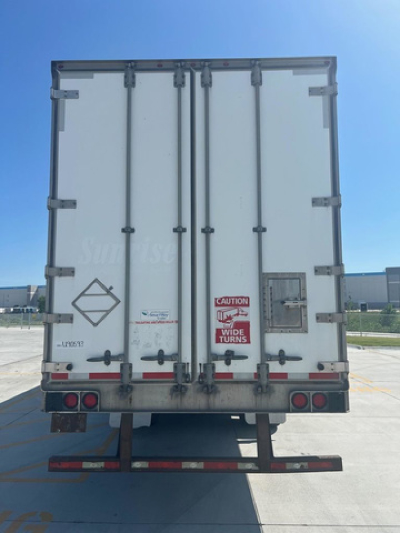 Back view of a white utility van trailer with double doors, red caution sticker, and taillights; licenses and markings along bottom edge.