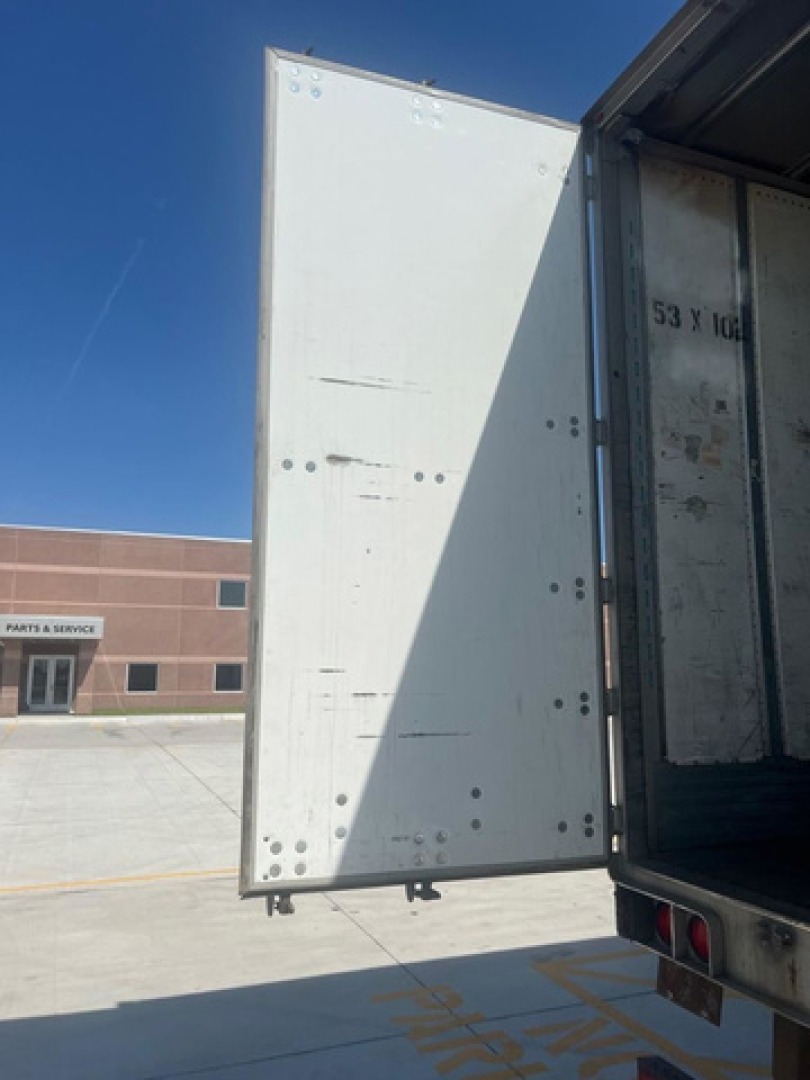 Trailer van rear door open showing interior with metal panel and latching hardware; exterior white panel with vertical seam, truck trailer rolling into loading dock area.