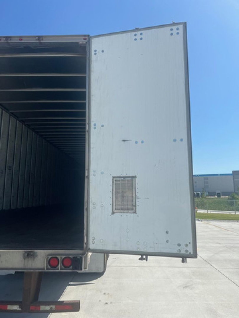 Truck trailer rear door open showing interior of cargo area and exterior door with small vent; white metal siding with rivets; blue sky.