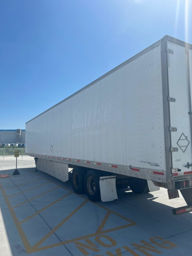 Utility van trailer in a parking lot with a long white semi-trailer, side view shows some faded branding and diamond placard at rear.