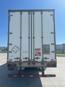 Back view of a white utility van trailer with double doors, red caution sticker, and taillights; licenses and markings along bottom edge.