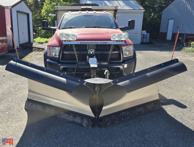 Ram 5500 front view with snow plow and sander attachment, black plow blades spread wide, red truck in background, dealership lot.