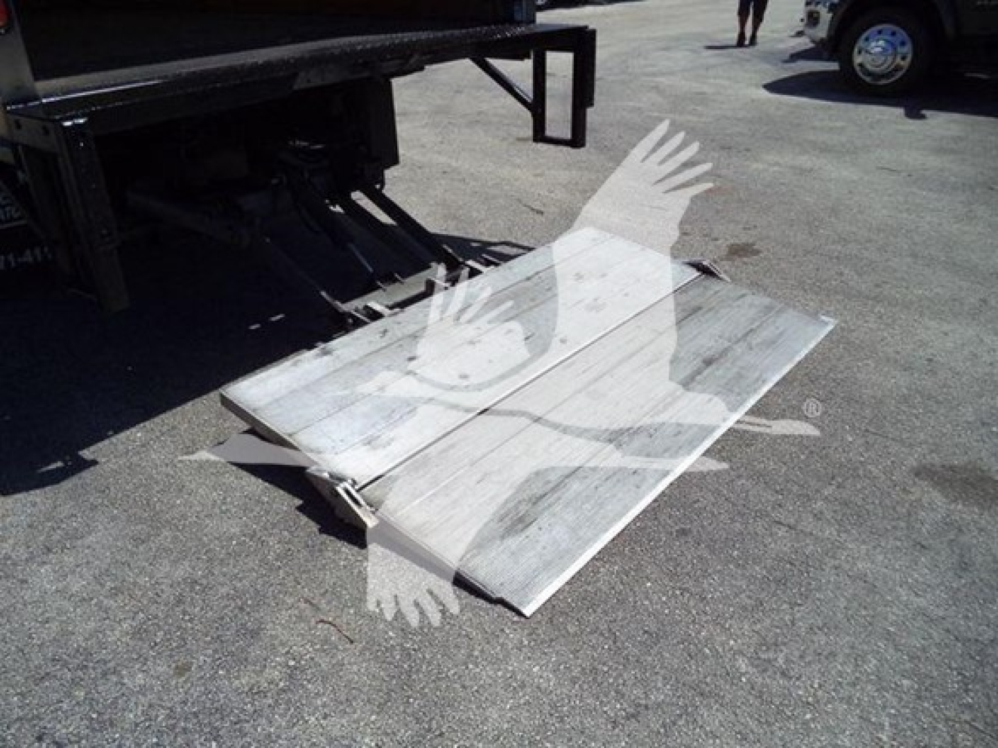 Isuzu NPR HD truck with a metal loading ramp or platform lowered from the back, showing a weathered aluminum surface and shadows on pavement.