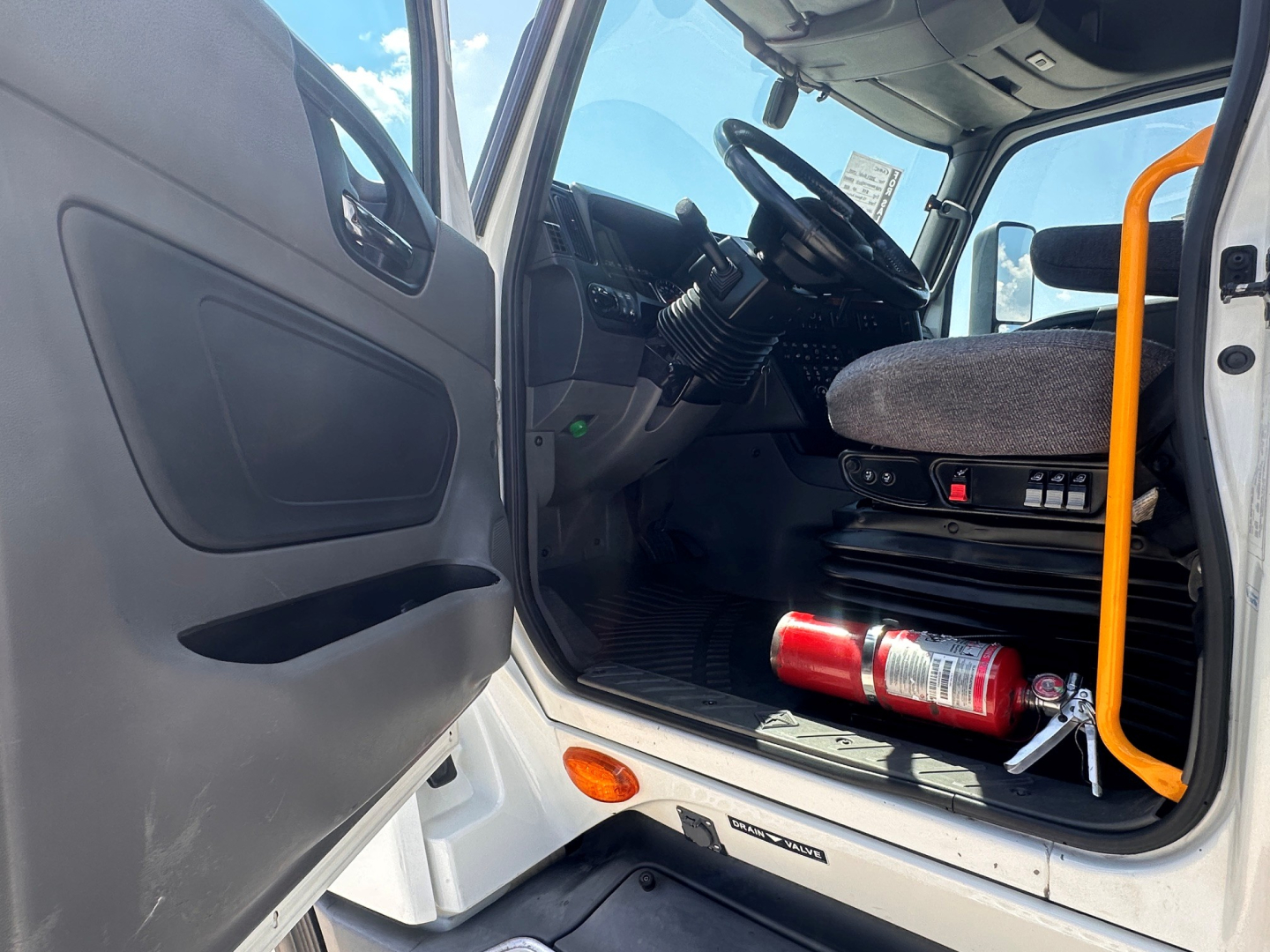 Truck cab interior of a 2021 International LT625: open driver door, steering wheel, dashboard, seat, fire extinguisher mounted under seat area, orange handrail visible.