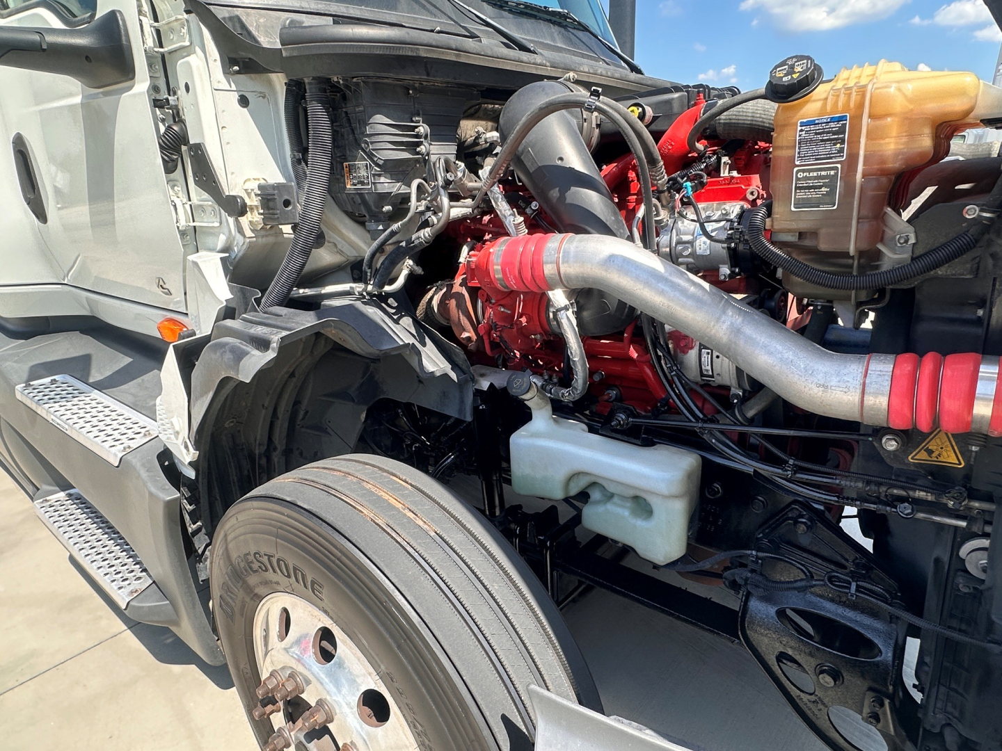 Truck engine bay of a 2021 International LT625 with visible coolant reservoir, intake piping, red engine block, and front tire, showing components and hose connections.